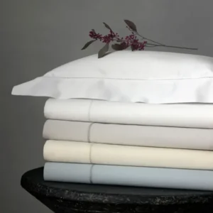 A stack of neatly folded bed linens in soft pastel colors, topped with a white pillow adorned with a sprig of purple flowers. Featuring Nocturne Hemstitch bedding by Matouk, available at Linen Society.