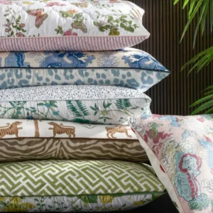 A stack of five decorative pillows with colorful patterns sits beside a sixth customizable Reversible Quilted Lumbar Pillow Cover by Matouk, with a green plant partially visible in the top right corner.
