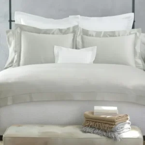 A neatly made bed with soft gray linens, multiple pillows, and decorative throws, set in a cozy bedroom with green plants nearby. Featuring Nocturne Hemstitch bedding by Matouk, available at Linen Society.