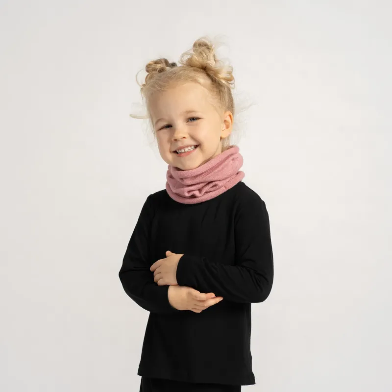 Little girl in a black outfit wearing a soft pink knit neck warmer, smiling with arms crossed.