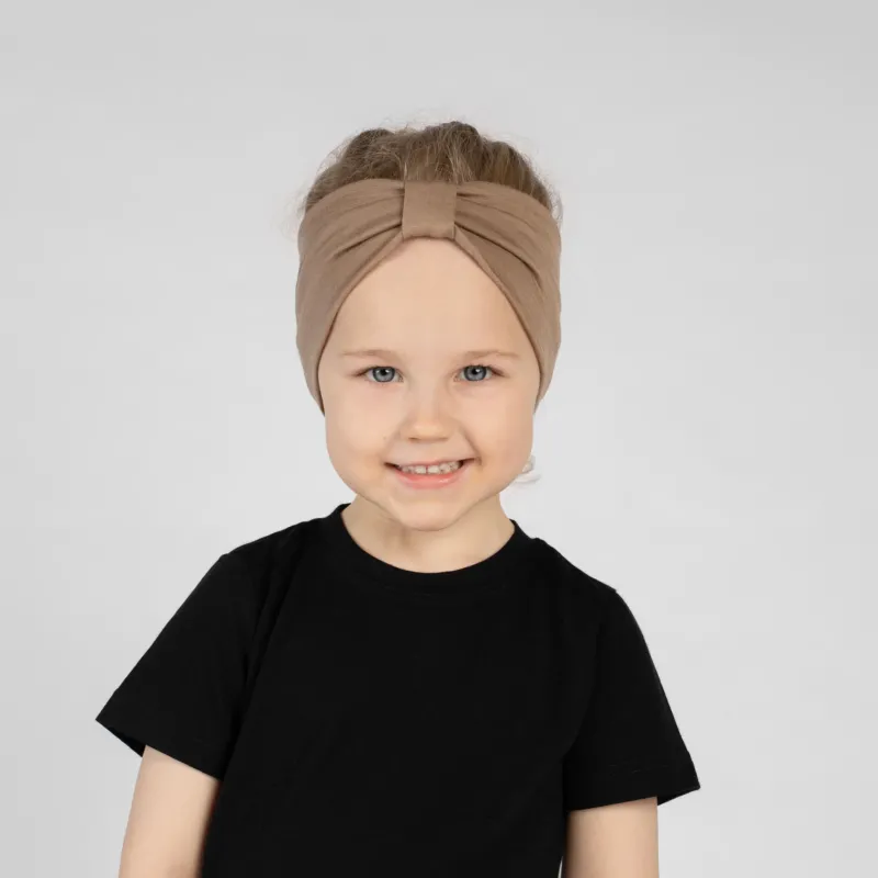 A close-up studio portrait of a young girl smiling directly at the camera against a plain light grey background. She is wearing a black short-sleeved t-shirt and a beige ribbon headband.