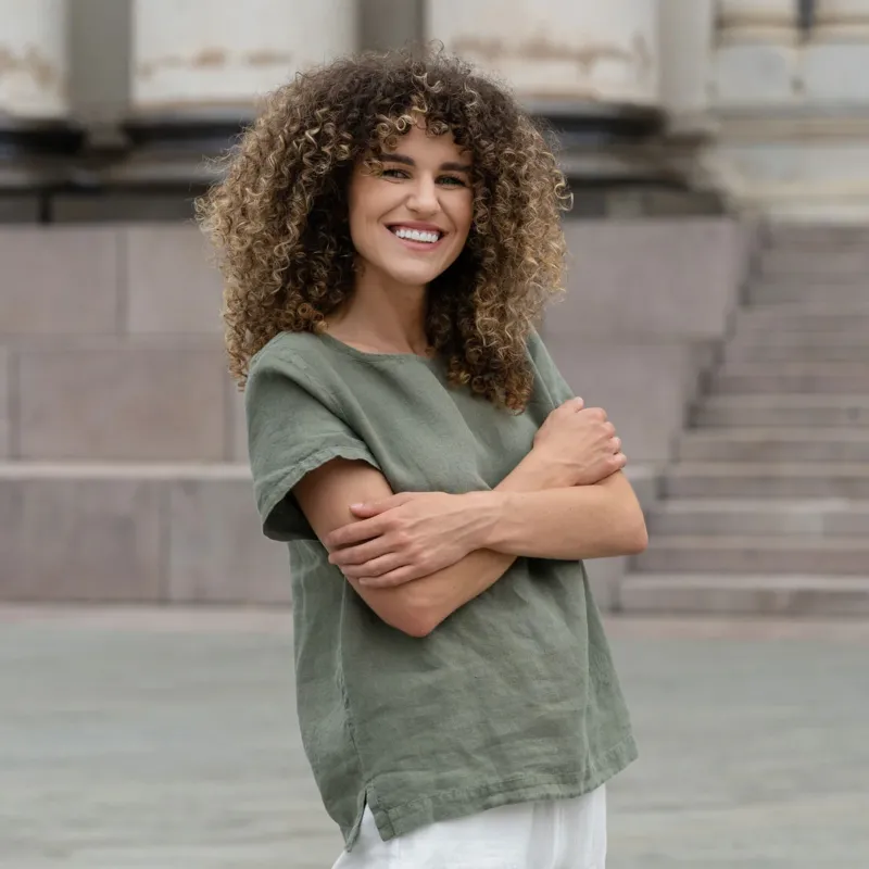 Beautiful woman with a curly hair in the city, posing wearing stone green linen blouse Emma and pure white linen pants Dakota.