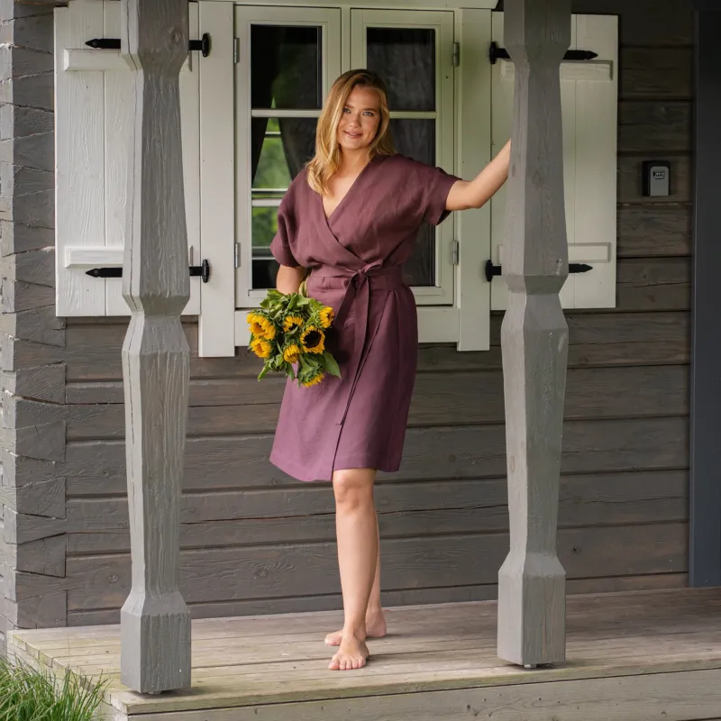 A barefoot woman poses on the wooden porch of a rustic house, resting one hand on a decorative pillar. She is wearing a knee-length, shadow purple linen wrap dress with a tie waist and holding a bouquet of fresh sunflowers.