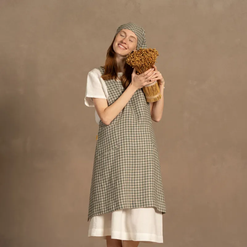 A person with fair skin and long reddish-brown hair stands smiling with closed eyes, gently holding a bundle of dried golden flowers to their chest. They are wearing a white short-sleeved linen shirt under a green check apron and a matching checkered headscarf tied under their chin. The background is a muted warm brown.