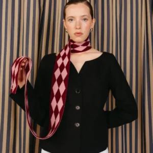 Model wearing a fitted black jacket and white pants, showcasing a TBCo Merino Wool Skinny Scarf in Burgundy Harlequin, elegantly draped around her neck.