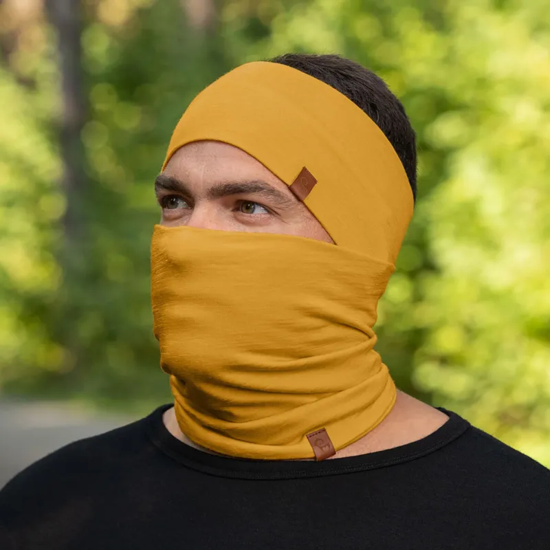 A person with short, dark hair is shown wearing a spicy yellow headband and a matching neck gaiter that is pulled up to cover their mouth and nose, with only their eyes visible. They are wearing a black long-sleeved shirt, and the blurred background suggests an outdoor setting with green foliage.