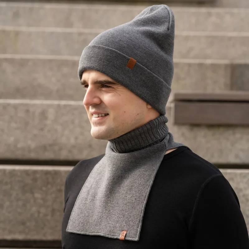 A man shown from the chest up, turned slightly to his right and smiling. He is wearing a dark grey knit beanie with a small brown logo patch and a matching dark grey knit neck warmer over a black long-sleeve shirt. The background consists of blurred concrete stairs.