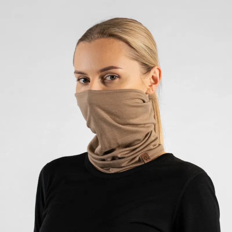 A studio portrait of a blonde woman looking directly at the camera. She is wearing a light beige neck gaiter pulled up to cover her nose and lower face, along with a solid black long-sleeved top. A small brown leather logo patch is visible on the bottom edge of the gaiter. The background is a plain light grey.