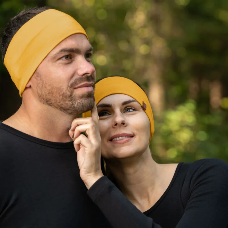 A man and a woman are closely positioned outdoors, both wearing spicy yellow menique merino wool headbands and black long-sleeved shirts. The man, on the left, has short brown hair and a beard and is looking upwards and to his left. The woman, on the right, has light brown hair and is looking upwards with a smile, her hand gently touching the man's face. The background is softly blurred with green and brown tones, suggesting a natural, possibly wooded, environment.