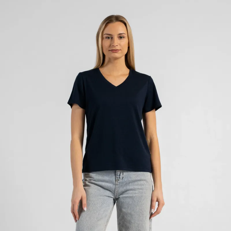 A studio portrait of a blonde woman standing and looking directly at the camera with her arms relaxed by her sides. She is wearing a regular fit, dark navy blue short-sleeved v-neck t-shirt and light-wash blue jeans. The background is a solid light grey.