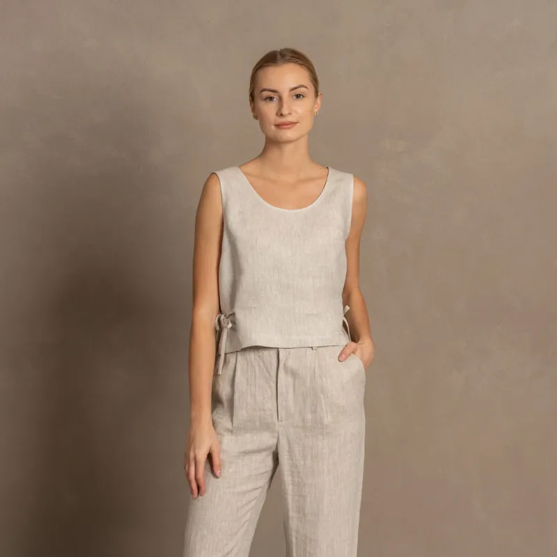 A woman with blonde hair pulled back wears a natural linen sleeveless crop top with side ties, paired with matching light natural linen pants. She stands facing forward with one hand in her pocket against a muted brown textured background.
