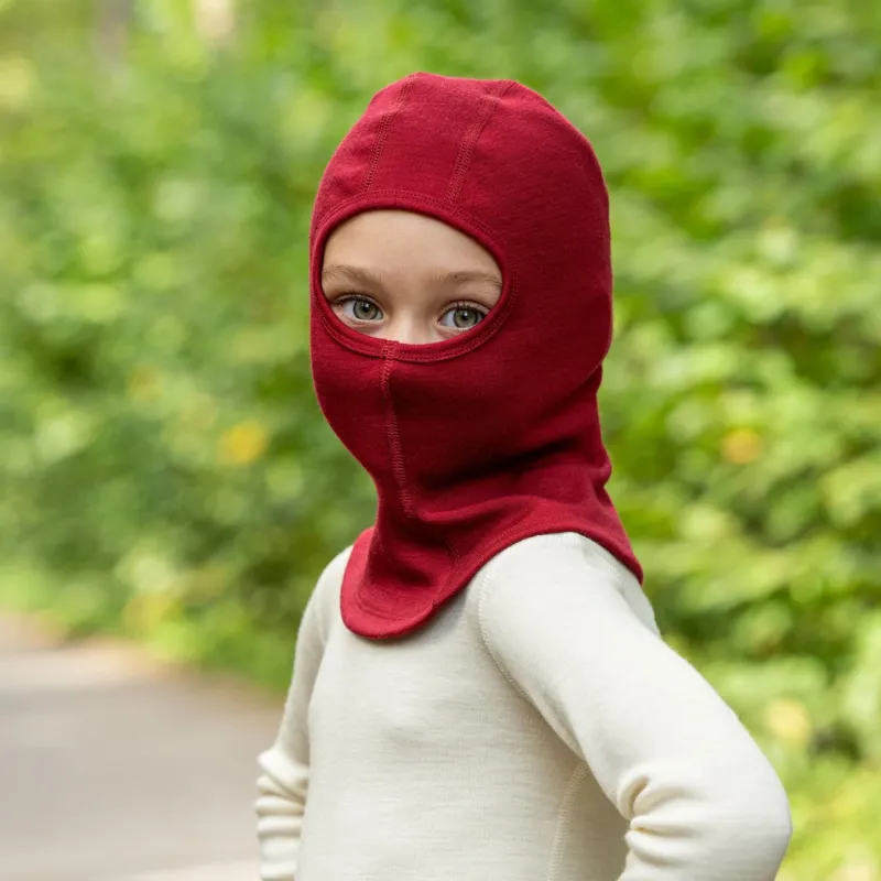 A young child wearing a royal cherry knit balaclava and a cream-colored shirt looks over their shoulder at the camera, with a background of blurred green foliage.